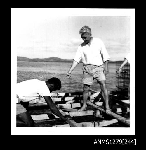 A man wearing a white shirt and shorts, and a Papua New Guinean man, on a wooden pearl production raft