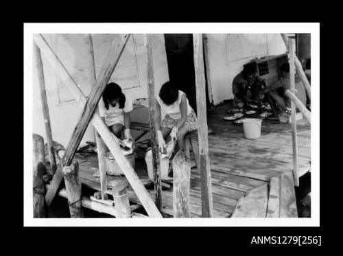 Two women scrubbing pearl shells over buckets, and men sitting in the background looking at pearl shells laid out on the ground, on a pearl raft