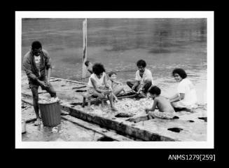 Photograph of men, women and children cleaning pearl shells, with a man on the left of the photograph discarding waste into a garbage bin