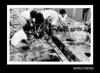 Photograph of men, women and children cleaning pearl shells, with three people crowded over a garbage bin