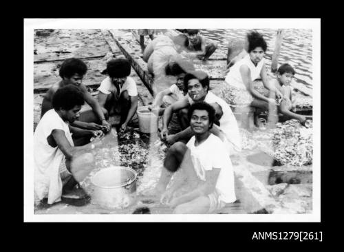 Photograph of men, women and children cleaning pearl shells, sitting around pearl shell cages