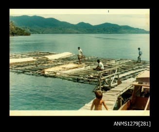 Colour photograph of three Papua New Guinean men standing or sitting on a wooden pearl production raft