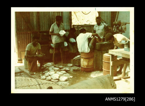 Colour photograph of several Papua New Guinean men and children in a shed, either using pearling equipment, holding pearl shells, or working with half pearls (or mabe pearls) in a metal cylinder