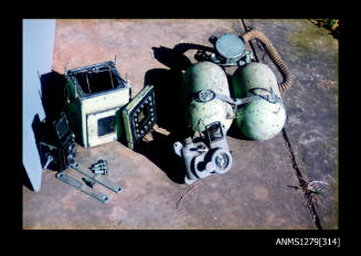 Colour photograph of a Rolleiflex camera, green underwater camera case, underwater camera and two green air tanks, sitting in a row on the cement