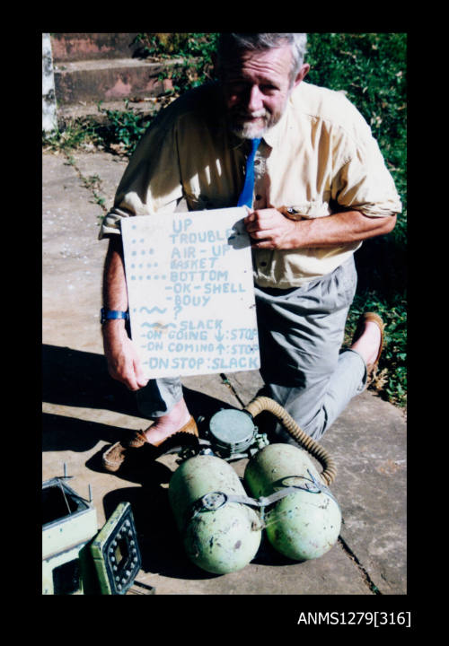 Colour photograph of a man holding a diving signal board, crounched dowm behind a green underwater camera case and green air tank