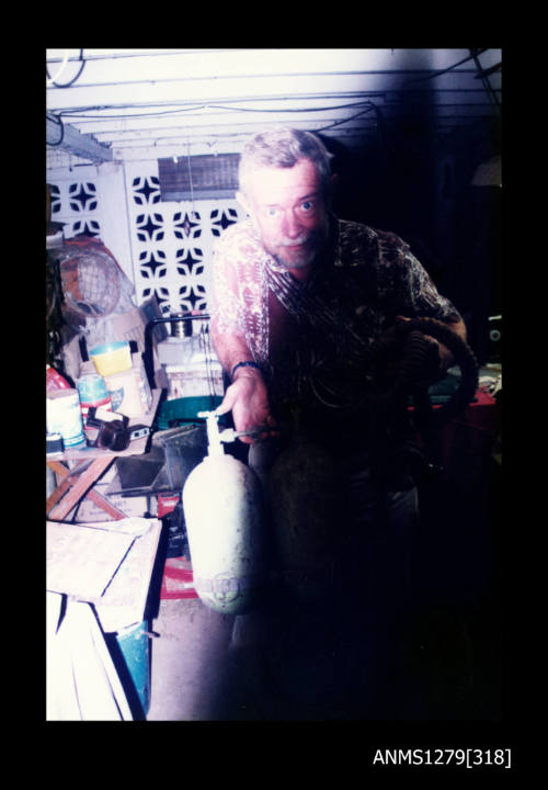 Colour photograph of a man standing underneath Denis George's house, holding two diving air tanks and diving hose