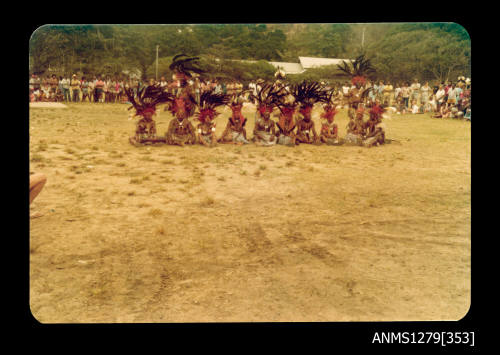 Colour photograph of several Papua New Guinean people in traditional dress, sitting in a row on the ground, surrounded by hundreds of onlookers, taken at the Pacific Festival on Pearl Island