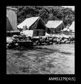 A rock wall on the beach, with tents, a shed and numerous cylindrical containers in the background