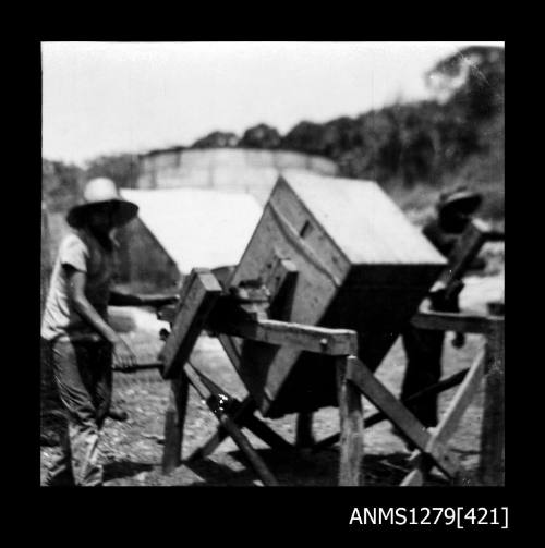 Photograph of man wearing a hat standing next to a wooden box on a stand, used to clean pearl cages