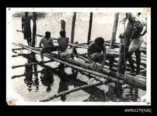 Three Papua New Guinean men standing on a beach, chopping long wooden logs