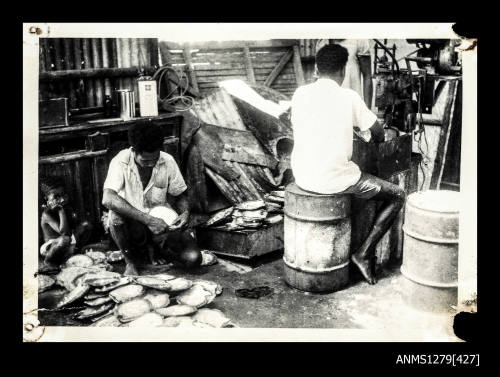 Two Papua New Guinean men and a child, with one of the men sitting beside a pile of pearl shells, and the other sitting on a metal cylinder at a machine