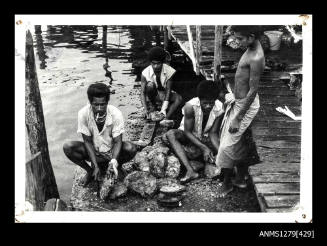 Photograph of four Papua New Guinean men, beside a wharf, opening pearl shells