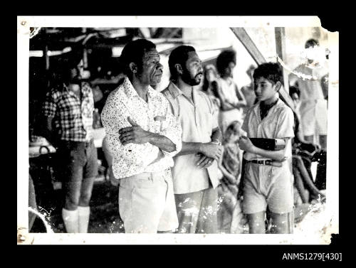 Photograph of several people, including two Papua New Guinean men and a child holding a box, looking into the distance