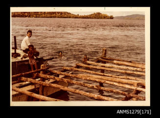 Colour photograph of a wooden pearl raft, with a man sitting on a boat with a young boy beside the raft, and Yurie (or Yulie) George and a young boy swimming in the water