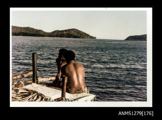Colour photograph of two Papua New Guinean men sitting on the edge of a boat, with landscape in the background