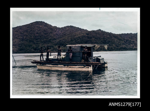 Colour photograph of a grey and cream coloured boat, with five men standing on the boat holding a wooden log