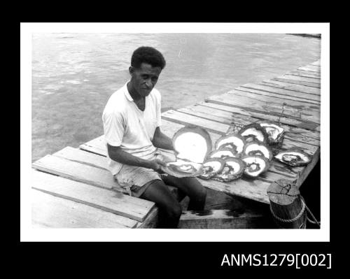 A Papua New Guinean man sitting on a wharf, holding an opened pearl shell, with pearl shells beside him on the wharf