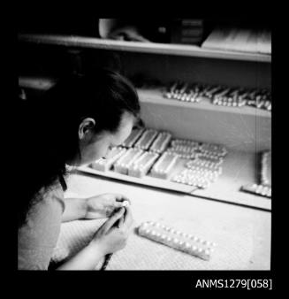 A woman holding a nucleus [?], at a desk containing wooden blocks with nuclei [?]