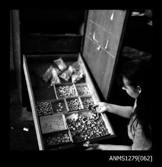 A woman holding out a draw with metal containers filled with half pearls (or mabe pearls), half pearl caps, and possibly pearl shell pieces