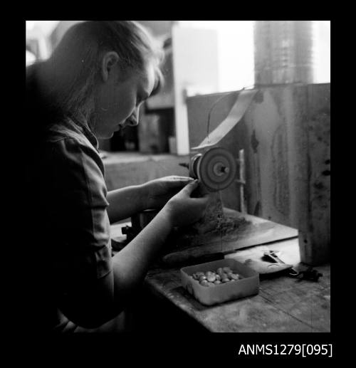 A woman sitting at a grinding machine, polishing or trimming half pearls (or mabe pearls) sitting in a container beside her