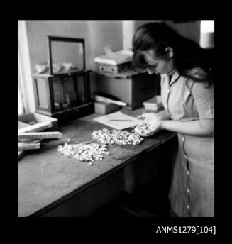 A woman standing next to a desk containing two piles of half pearls (or mabe pearls), with some also in her hands