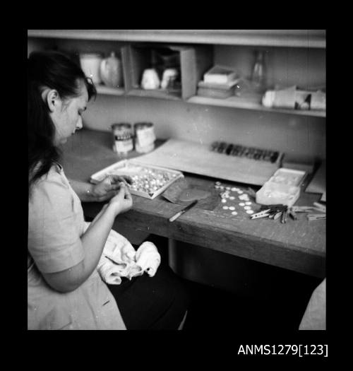 A woman sitting at a work desk with a large metal container filled with half pearls (or mabe pearls), and pearl producing equipment