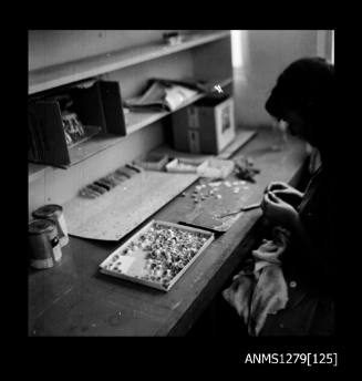 A woman sitting at a work desk with a large metal container filled with half pearls (or mabe pearls), and pearl producing equipment