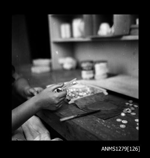 A woman sitting at a desk containing a tray of half pearls (or mabe pearls), holding a peg