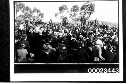 Crowd of people gathered underneath a row of flags with trees and a building in the background