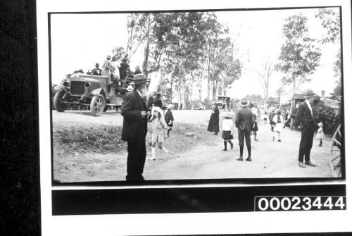 A crowd of people around a dirt road and embankment with two motor vehicles in the background