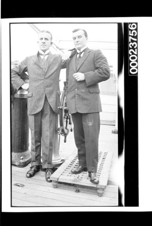 Two men wearing suits standing close to ships wheel