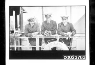 Three young officers in dark uniforms, are facing camera and leaning on the ship's rail, lifebuoy in front reads SS ITAURA