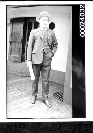 Man standing on deck wearing a a suit and boater and holding papers