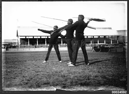 Three Indigenous Australian men holding spears in a grass paddock