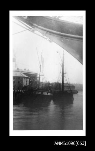 Black and white photograph of two vessels side by side at jetty to left with sterns facing camera