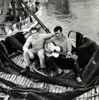 Photograph of two men sitting in a boat with nets on the floor, one playing guitar