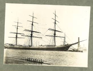 Full-rigged ship docked at a port, cox rowing eight in foreground