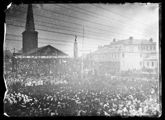 A crowd at the unveiling of a statue in Queens Square, Sydney with a church building and a banner reading 'God save the Queen' visible
