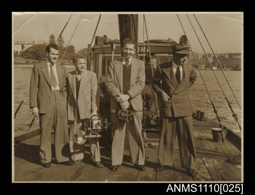 Photograph of four men in suits standing on the deck of a boat