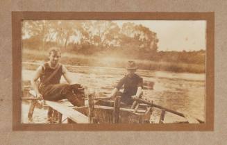 Two men by a river, one sitting in a dinghy with oars outstretched