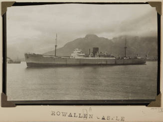 Photograph of  ROWALLAN CASTLE depicting port side of cargo ship under way off shore with cloud shrouded mountains off starboard side