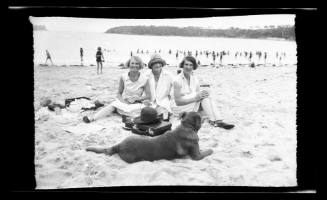 Three women sitting with a dog on a busy beach