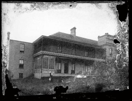 Large two storey house at unknown location with a woman standing at the base of the verandah