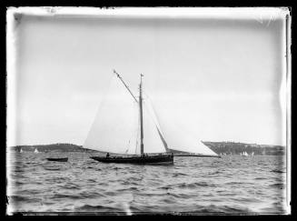 Gaff-rigged cutter with extended bow-sprit and one crew visible sails on Sydney Harbour with Vaucluse and Rose Bay in distance