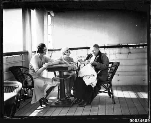 Three women seated around a table on the deck of the SS ORMISTON, one of them doing needlework