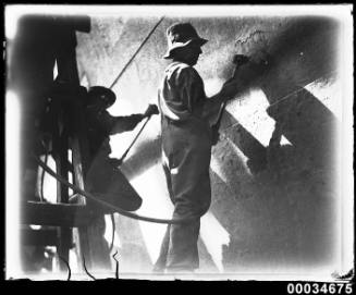 Two men scraping rust from the hull of a ship, possibly French warship BELLATRIX