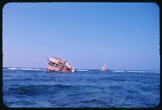 The front part of a shipwreck sitting on its side and a ship approaching in the distance