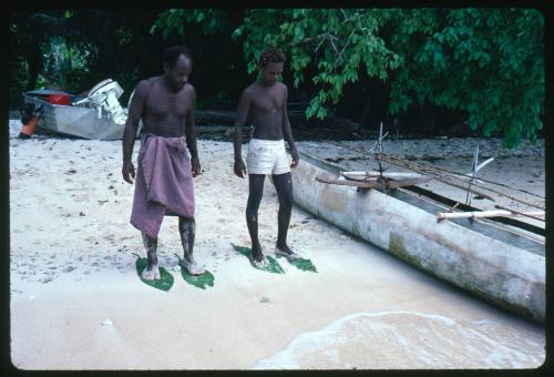 Two men on standing on beach next to outrigger canoe