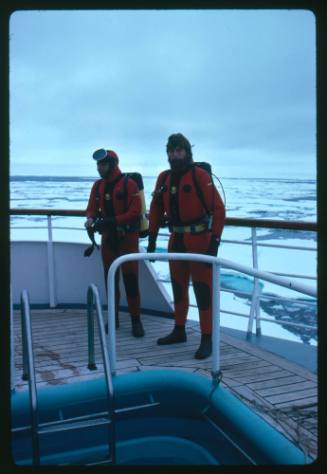 Two men in red diving suits on the deck of a vessel