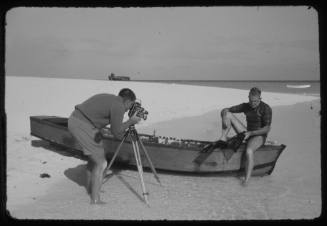 Black and white photos of a man filming a diver putting on fins while sitting on the edge of a small boat at the beach